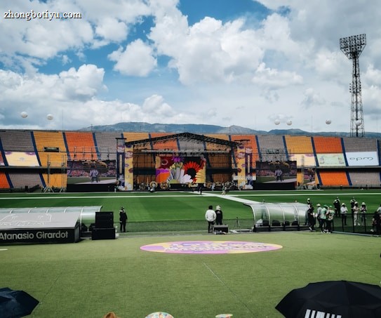 A sports stadium with a large stage set up on the field, hosting an outdoor event. Spectators wearing colorful hats sit in the foreground. The seating areas display vibrant colors, and a clear blue sky with scattered clouds is visible. Large screens display graphics related to the event.