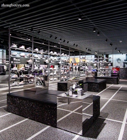 A modern sneaker store with sleek black shelving displaying various athletic shoes and sports apparel. Central seating areas and spotlights provide a contemporary retail ambiance. The signage prominently features a famous sportswear slogan in the background.