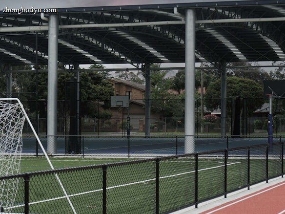 An outdoor sports facility featuring a running track and a soccer goal. The track is red, surrounded by a black metal fence, and the field is covered with artificial green turf. In the background, a basketball court is visible under a covered roof with metal supports. The sky is overcast, adding a moody atmosphere.