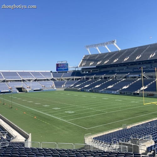 A large, empty sports stadium with blue seats surrounding a well-maintained green grass field. Bright sunlight creates clear shadows, and there are two goal posts on opposite ends of the field. A big electronic scoreboard displays some graphics, and the sky is clear and blue.